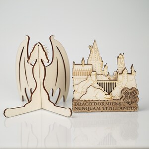 May include: A wooden model of Hogwarts castle with a dragon sculpture. The castle is detailed with windows and towers. The dragon is standing on a base with the text "Draco Dormiens Nunquam Titillandus".