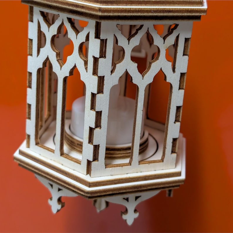 May include: A decorative wooden lantern with a white finish and intricate cut-out designs. The lantern features a candle inside, creating a warm glow. The lantern has a hexagonal base and decorative feet.