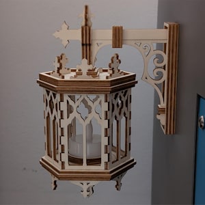 May include: A wooden wall-mounted lantern with an ornate, gothic design. The lantern features a hexagonal body with decorative cutouts and a small candle inside. The bracket and cross-shaped top are also made of wood, creating a warm, natural aesthetic.