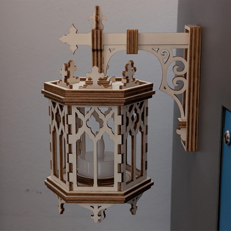May include: A wooden wall-mounted lantern with an ornate, gothic design. The lantern features a hexagonal body with decorative cutouts and a small candle inside. The bracket and cross-shaped top are also made of wood, creating a warm, natural aesthetic.
