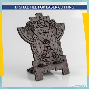 Può includere: Un supporto in legno tagliato al laser a tema vichingo. Il supporto presenta un design a scudo con asce, rune e un nodo celtico. Il testo "DIGITAL FILE FOR LASER CUTTING" è in alto. Il supporto è grigio e ha una trama di venature del legno.
