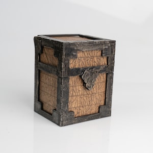May include: A small, square wooden chest with dark metal accents. The chest features a textured wood grain pattern and is secured with a decorative metal clasp. The corners and edges are reinforced with dark metal.