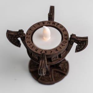 Pu&ograve; includere: Un portacandele in legno a tema vichingo. Il supporto marrone presenta due decorazioni a forma di ascia e un design circolare con rune. Una candela LED bianca &egrave; al centro, che fornisce una luce soffusa. La base ha un motivo decorativo.
