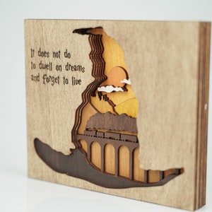 May include: Wooden wall art featuring a 3D layered design of a train traveling over a bridge with a sunset in the background. The quote "It does not do to dwell on dreams and forget to live" is engraved on the left side of the art.