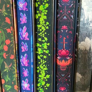 May include: A selection of decorative book spines with diverse designs. One spine showcases purple crystal formations on a blue backdrop. Another features green ivy on a black background. Other spines display floral and fantasy-themed artwork. The spines are vertical and closely aligned.