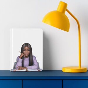 May include: A white notebook with a purple-clad woman with long black hair sitting at a desk with a pen in her hand. The notebook has a pink and white striped design on the inside cover.