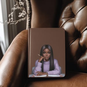 May include: A brown leather-bound journal with a colorful illustration of a woman with long black hair, wearing a purple sweater, sitting at a desk with a pen in her hand.
