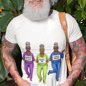 Puede incluir: Camiseta blanca con un dise&ntilde;o humor&iacute;stico de tres mujeres mayores con ch&aacute;ndales. El texto de la camiseta dice "Leader of the Pack", "Wasn't Me" y "Caught Smiling".