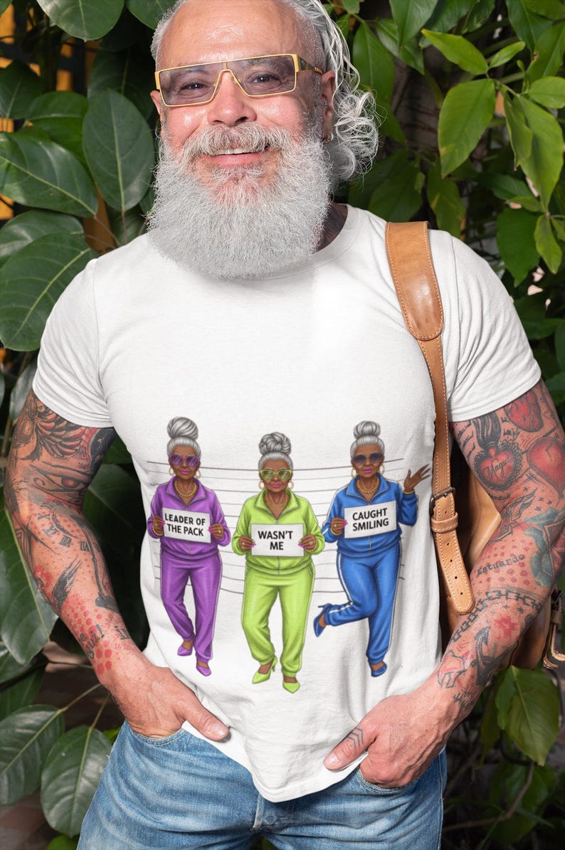 Puede incluir: Camiseta blanca con un dise&ntilde;o humor&iacute;stico de tres mujeres mayores con ch&aacute;ndales. El texto de la camiseta dice "Leader of the Pack", "Wasn't Me" y "Caught Smiling".