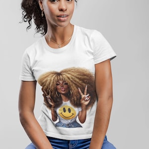 Puede incluir: Una camiseta blanca con un gr&aacute;fico de una mujer con pelo rizado grande, haciendo el signo de la paz y con una camiseta de carita sonriente. La mujer lleva vaqueros azules y joyas doradas. El fondo es gris claro.