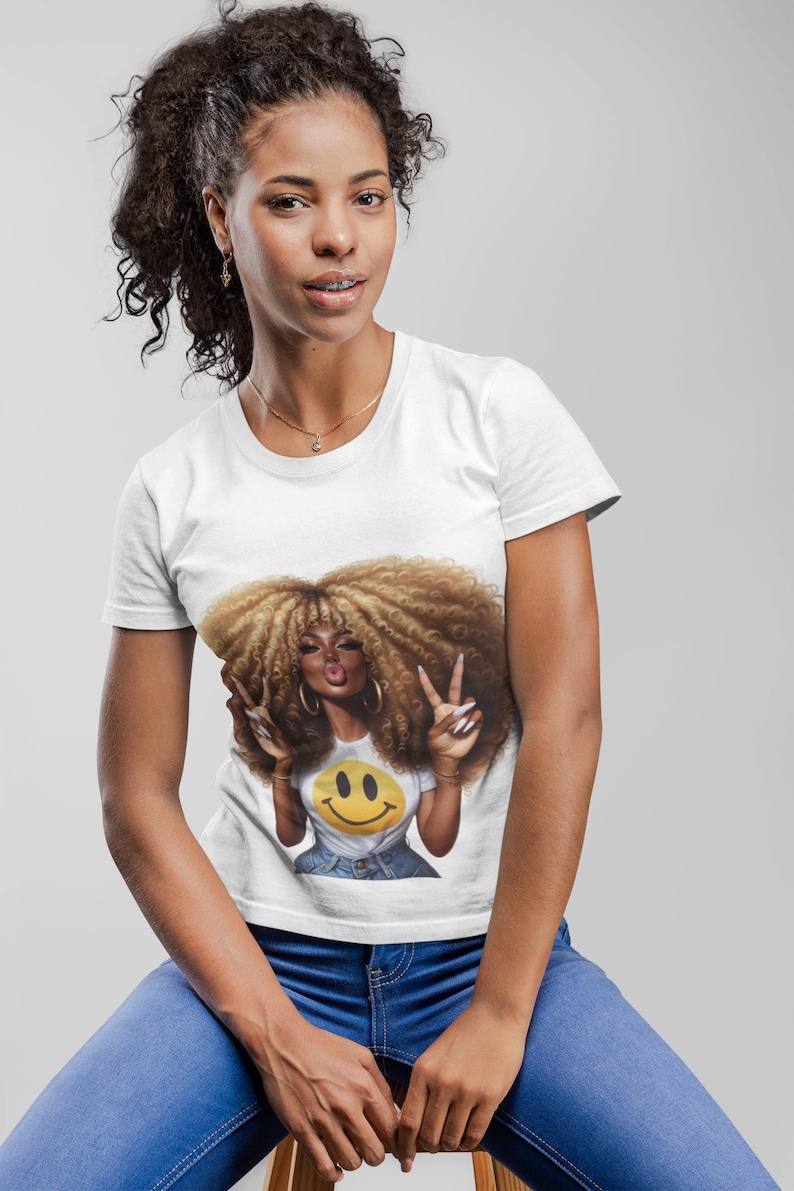Puede incluir: Una camiseta blanca con un gr&aacute;fico de una mujer con pelo rizado grande, haciendo el signo de la paz y con una camiseta de carita sonriente. La mujer lleva vaqueros azules y joyas doradas. El fondo es gris claro.