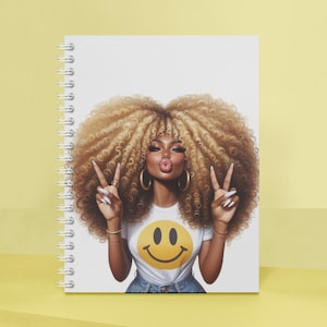 Puede incluir: Cuaderno de espiral con una colorida ilustraci&oacute;n de una mujer con cabello rubio voluminoso, haciendo el signo de la paz. Lleva una camiseta blanca con un emoji amarillo. El cuaderno tiene una cubierta blanca.