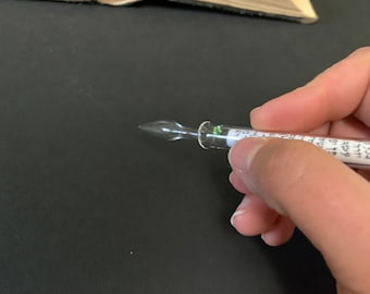 May include: A clear glass calligraphy pen with a pointed tip and a label with handwritten text. The pen is held in a hand.