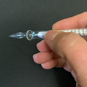 Może przedstawiać: Pióro stalowe z przezroczystego szkła z niebieskim i zielonym kamieniem szlachetnym na górze. Pióro jest trzymane w dłoni, a korpus jest wypełniony zwiniętym kawałkiem papieru.