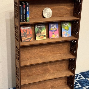 Puede incluir: Una estantería de madera en miniatura con cinco estantes. El estante superior tiene una pequeña moneda encima. El segundo estante tiene cinco libros de bolsillo, incluyendo Goosebumps, Night of the Living Dummy, Matilda y The BFG. Los estantes restantes están vacíos.