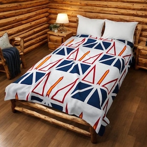 May include: A white blanket with a geometric pattern of blue triangles, red triangles, and orange lines. The blanket is on a wooden bed frame in a rustic cabin setting. Two white pillows are on the bed.