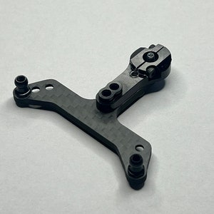 Op de afbeelding: Een zwart koolstofvezel onderdeel met meerdere bevestigingspunten en een draaibaar gedeelte. Het onderdeel heeft een getextureerd oppervlak en lijkt voor een mechanisch apparaat of model te zijn. Het onderdeel is ontworpen met precisie en duurzaamheid.