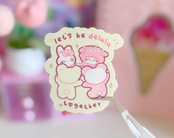 Puede incluir: Una pegatina de color pastel con dos personajes de dibujos animados, un conejo y un oso, tomados de la mano. El texto "let's be delulu" y "together" está escrito en la pegatina. La pegatina está sujeta con unas pinzas.