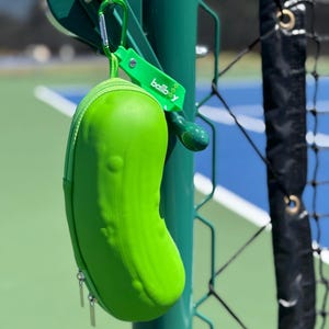 K&ouml;nnte beinhalten: Ein leuchtend gr&uuml;ner, picklef&ouml;rmiger Tennisballbeh&auml;lter mit Rei&szlig;verschluss. Der Beh&auml;lter h&auml;ngt an einem gr&uuml;nen Metallzaunpfosten mit einem Karabinerhaken und einem gr&uuml;nen Etikett mit der Aufschrift "ballboy".