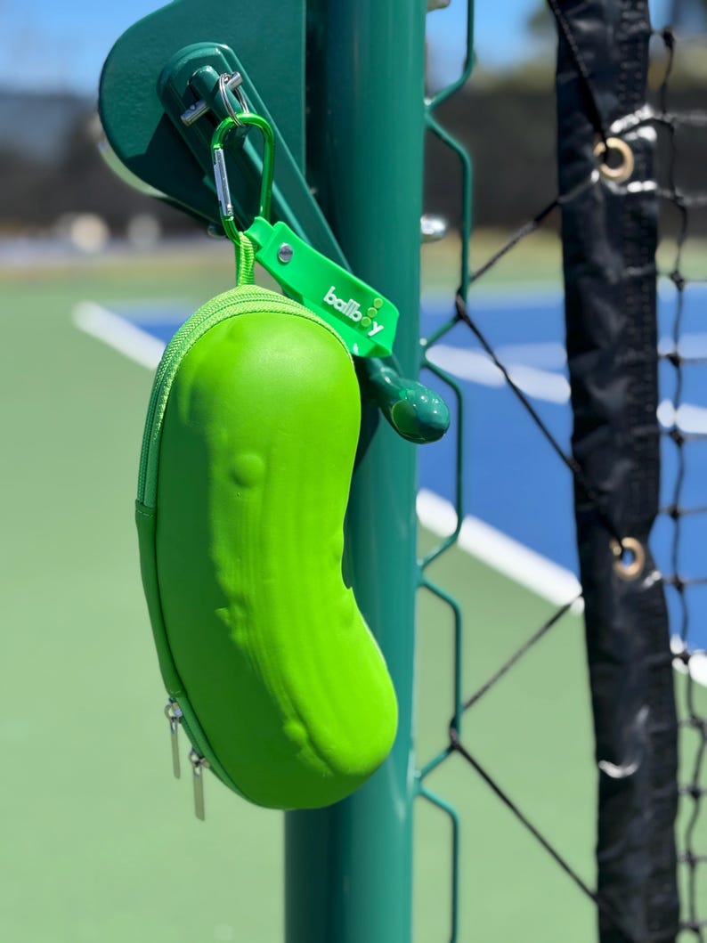 K&ouml;nnte beinhalten: Ein leuchtend gr&uuml;ner, picklef&ouml;rmiger Tennisballbeh&auml;lter mit Rei&szlig;verschluss. Der Beh&auml;lter h&auml;ngt an einem gr&uuml;nen Metallzaunpfosten mit einem Karabinerhaken und einem gr&uuml;nen Etikett mit der Aufschrift "ballboy".