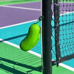 K&ouml;nnte beinhalten: Ein gr&uuml;ner, gurkenf&ouml;rmiger Beh&auml;lter, der an einem schwarzen Metallpfosten mit einem wei&szlig;en Netz dahinter h&auml;ngt. Der Beh&auml;lter h&auml;ngt an einem Tennisplatz.