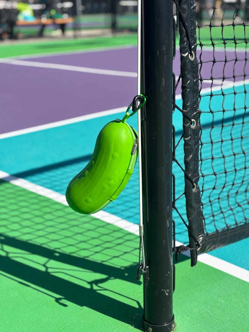 K&ouml;nnte beinhalten: Ein gr&uuml;ner, gurkenf&ouml;rmiger Beh&auml;lter, der an einem schwarzen Metallpfosten mit einem wei&szlig;en Netz dahinter h&auml;ngt. Der Beh&auml;lter h&auml;ngt an einem Tennisplatz.