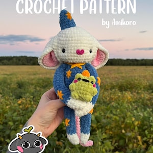 May include: A crochet pattern by Amikoro featuring a plush toy with a white head, large ears with pink inner details, and a blue body with yellow star accents. The toy wears a matching blue hat with a yellow star and holds a small green frog plush.