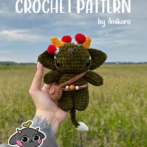 Puede incluir: Una criatura de peluche tejida a ganchillo en color verde oscuro con orejas grandes, cuernos naranjas y pompones rojos. Lleva un bolso marrón y un cinturón con cuentas de madera. El texto CROCHET PATTERN by Amikoro aparece en la parte superior.