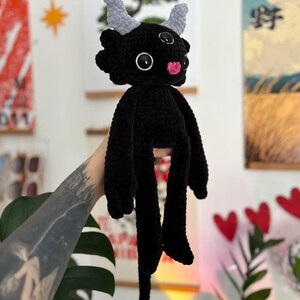 May include: A black crocheted plush creature featuring two light gray horns, three black and white eyes, and a small pink nose. The figure has long arms, long legs, and a thin tail ending in a light gray heart-shaped tip, all crafted from yarn.