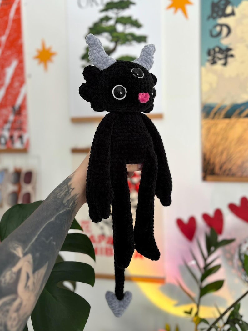 May include: A black crocheted plush creature featuring two light gray horns, three black and white eyes, and a small pink nose. The figure has long arms, long legs, and a thin tail ending in a light gray heart-shaped tip, all crafted from yarn.