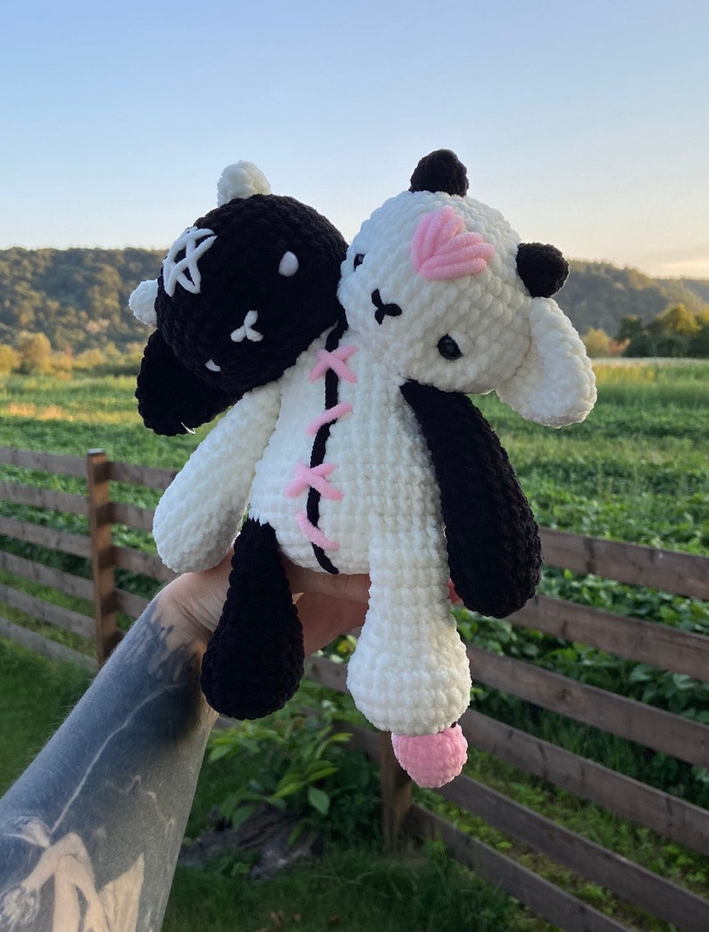 May include: A handmade, two-headed plush toy. One head is black with a white star, the other is white with a pink heart. The toy is stitched together with pink yarn and has black and white limbs.