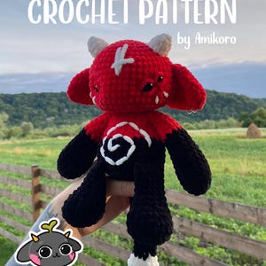 Puede incluir: Un peluche de ganchillo rojo y negro con detalles blancos, incluyendo cuernos y una cruz en la cabeza. El texto "CROCHET PATTERN" y "by Amikoro" es visible. El fondo es borroso, con campos y árboles.