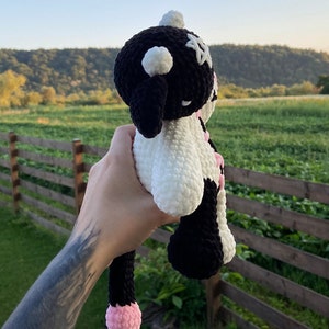 May include: A handmade crochet cow plushie in black and white with pink accents. The plushie has a round head with black ears, a white face, and a long black tail with a pink tip. The plushie is held up against a backdrop of a green field and a wooden fence.