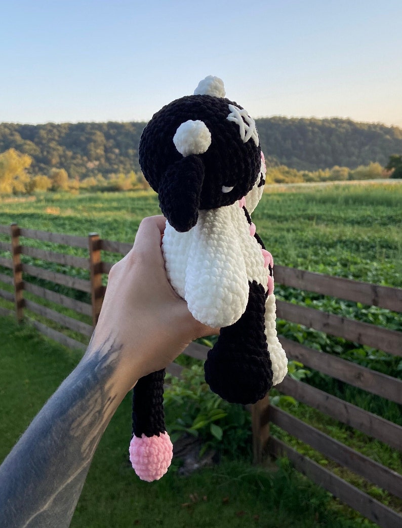 May include: A handmade crochet cow plushie in black and white with pink accents. The plushie has a round head with black ears, a white face, and a long black tail with a pink tip. The plushie is held up against a backdrop of a green field and a wooden fence.