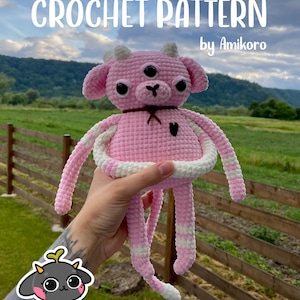 May include: A pink crochet creature with three eyes, small horns, and long limbs. The text "CROCHET PATTERN" and "by Amikoro" are displayed at the top. The creature has a black heart and a brown bow. The background shows a green field and a wooden fence.