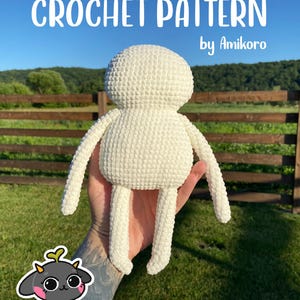 Puede incluir: Una muñeca de ganchillo de color crema con brazos y piernas largos se sostiene en una mano. El texto "CROCHET PATTERN" y "by Amikoro" se muestran encima de la muñeca. El fondo presenta una valla de madera, hierba verde y un cielo azul.