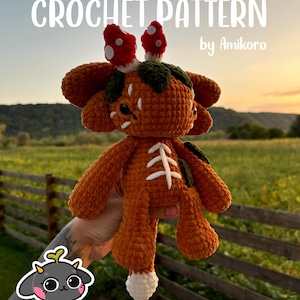 May include: A crocheted orange creature with white detailing and red mushroom-shaped headwear. The text "CROCHET PATTERN by Amikoro" is displayed at the top. The creature is held in front of a blurred outdoor background.