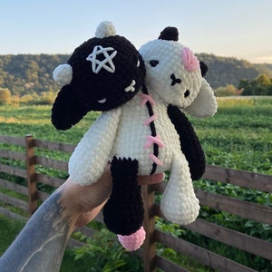 May include: A handmade, two-headed plush toy. One head is black with a white star, the other is white with pink accents. The body is white and black, with pink stitching. The toy is held up against a backdrop of green fields and a blue sky.