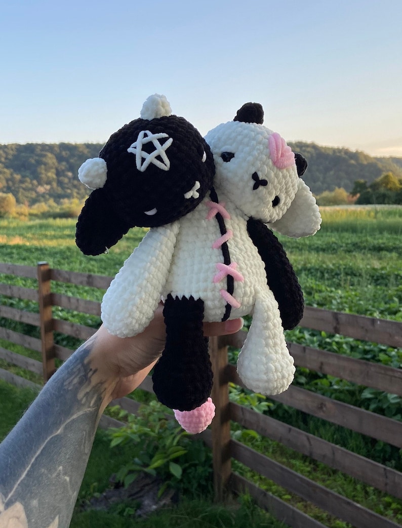 May include: A handmade, two-headed plush toy. One head is black with a white star, the other is white with pink accents. The body is white and black, with pink stitching. The toy is held up against a backdrop of green fields and a blue sky.