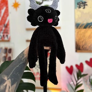 May include: A black crocheted plush creature featuring three eyes with black pupils, two light gray textured horns, and a small pink tongue. The creature has long arms, a long tail ending in a light gray heart-shaped tip, and a soft, fuzzy texture.