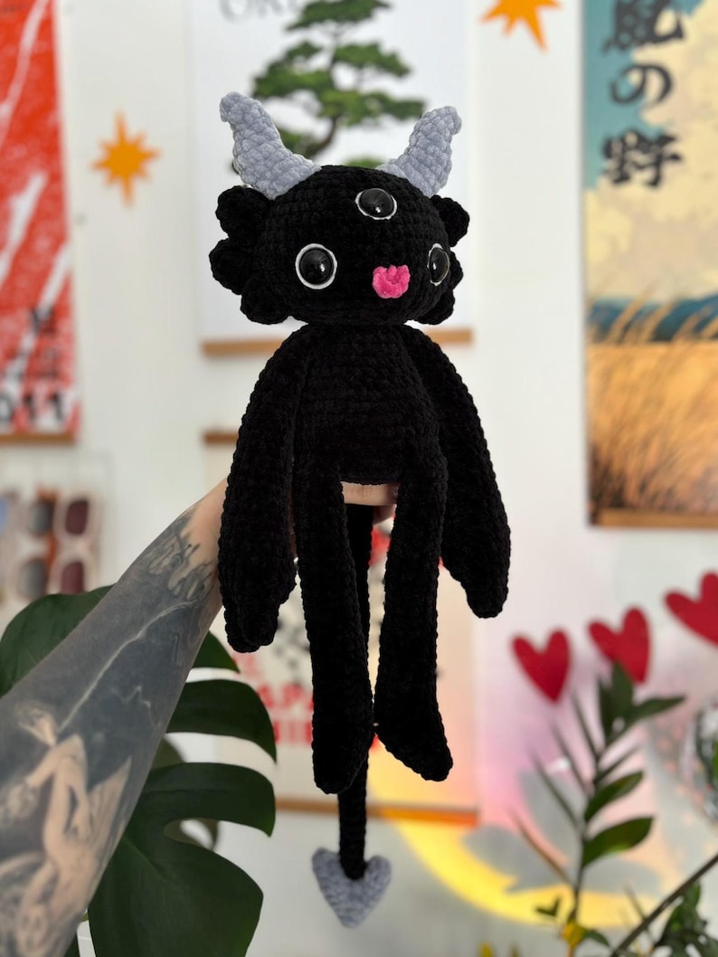 May include: A black crocheted plush creature featuring three eyes with black pupils, two light gray textured horns, and a small pink tongue. The creature has long arms, a long tail ending in a light gray heart-shaped tip, and a soft, fuzzy texture.