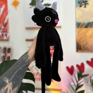 May include: A black crocheted plush toy featuring two light gray horns, large black button eyes with white centers, and a small pink tongue. The creature has long arms, a long tail ending in a gray arrow-shaped tip, and a textured yarn finish.