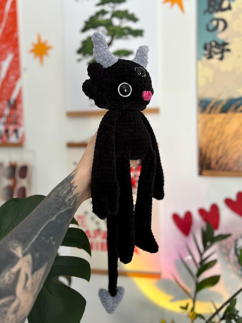 May include: A black crocheted plush toy featuring two light gray horns, large black button eyes with white centers, and a small pink tongue. The creature has long arms, a long tail ending in a gray arrow-shaped tip, and a textured yarn finish.