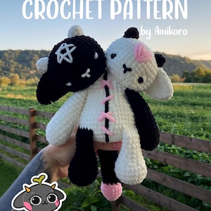 May include: A handmade crochet pattern for a plush toy, featuring a black and white sheep with pink accents. The text "CROCHET PATTERN by Amikoro" is displayed at the top. The toy is held in front of a green field and a wooden fence.