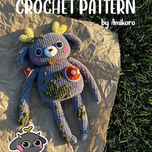 Op de afbeelding: Een gehaakte hert knuffel met gele geweien, een bruine kraag en een paddenstoel accent. De knuffel is grijs met groene en oranje details. De afbeelding bevat de tekst "CROCHET PATTERN by Amikoro" in witte letters.