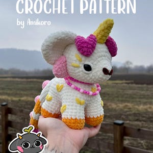 May include: A handmade crochet unicorn toy with a white body, pink and yellow accents, and a yellow horn. The toy has a pink beaded necklace and is held in a person's hand. The text "CROCHET PATTERN by Amikoro" is at the top of the image.
