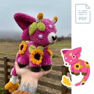 Könnte beinhalten: Eine rosa gehäkelte Amigurumi-Spielzeugkuh mit einem langen Schwanz, grünen und gelben Flecken und einem Sonnenblumenmuster. Die Kuh trägt eine silberne Kettenkette und hält eine Sonnenblume im Maul.
