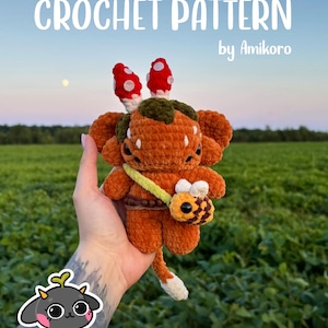 May include: A crochet pattern by Amikoro featuring a plush creature with orange yarn, two red and white mushroom-like protrusions on its head, a dark green patch, and a small yellow and brown bee-shaped bag worn across its body with a long tail.
