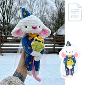 May include: A white crocheted mouse wearing a blue and gold star-patterned outfit and a pointed hat holds a green crocheted frog. The mouse has pink ears and a yellow ribbon around its neck. The mouse is holding the frog in its front paws.