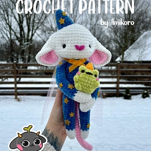 Może przedstawiać: Szydełkowa mysz w niebieskim stroju czarodzieja ze złotymi gwiazdami i pasującym kapeluszem. Mysz trzyma zieloną żabę. Tekst "CROCHET PATTERN" i "by Amikoro" jest widoczny na zdjęciu.